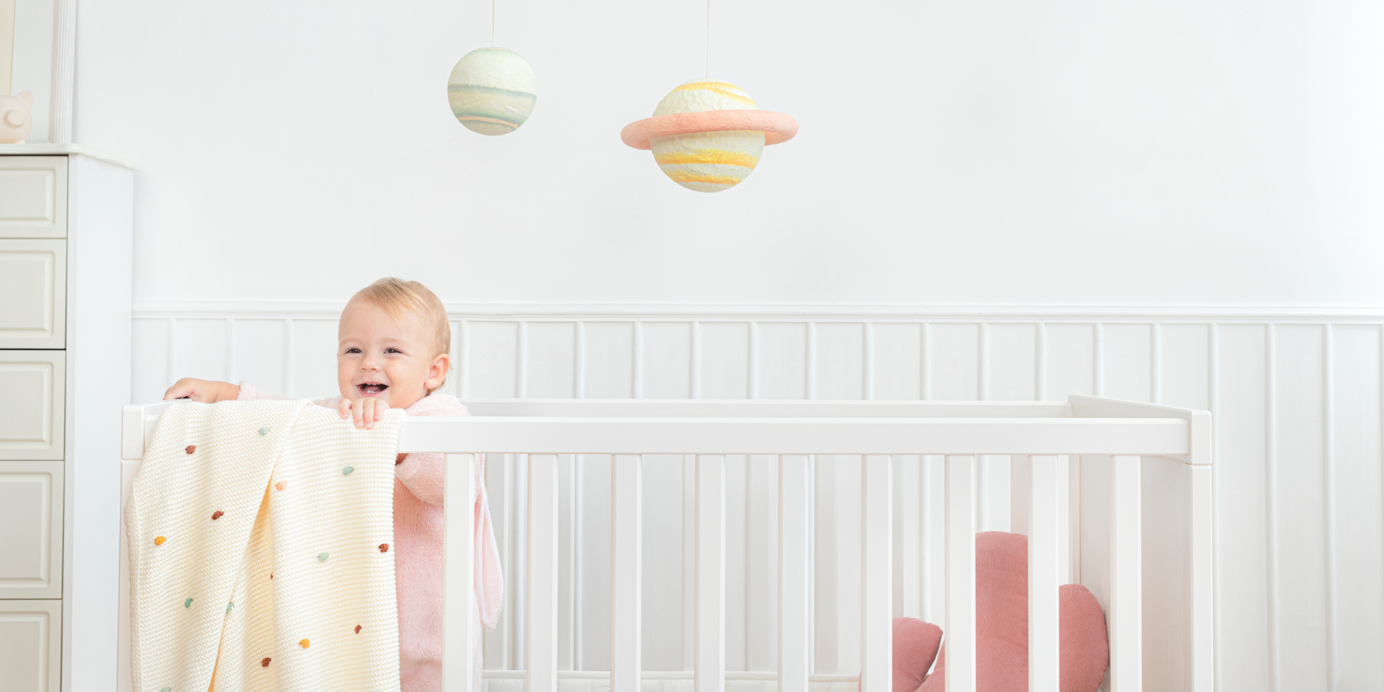 Baby in a crib with a white blanket and colorful hanging toys.

