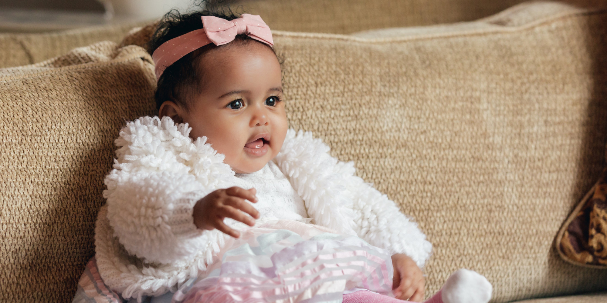 Baby in a white outfit with a pink bow sitting on a beige couch.

